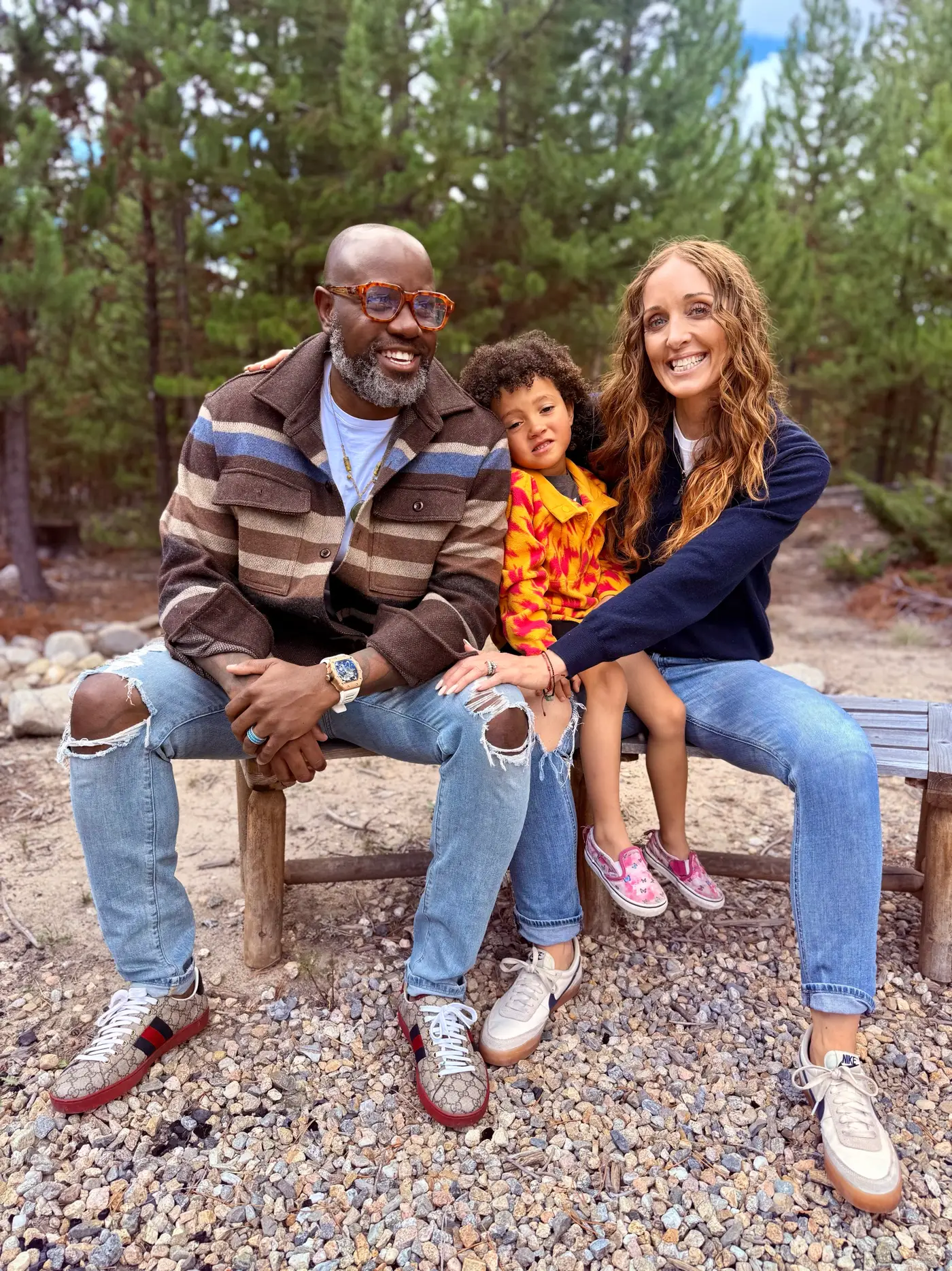 Hugo and Shannon “Redd” sitting on a bench in a pine forest with their young child