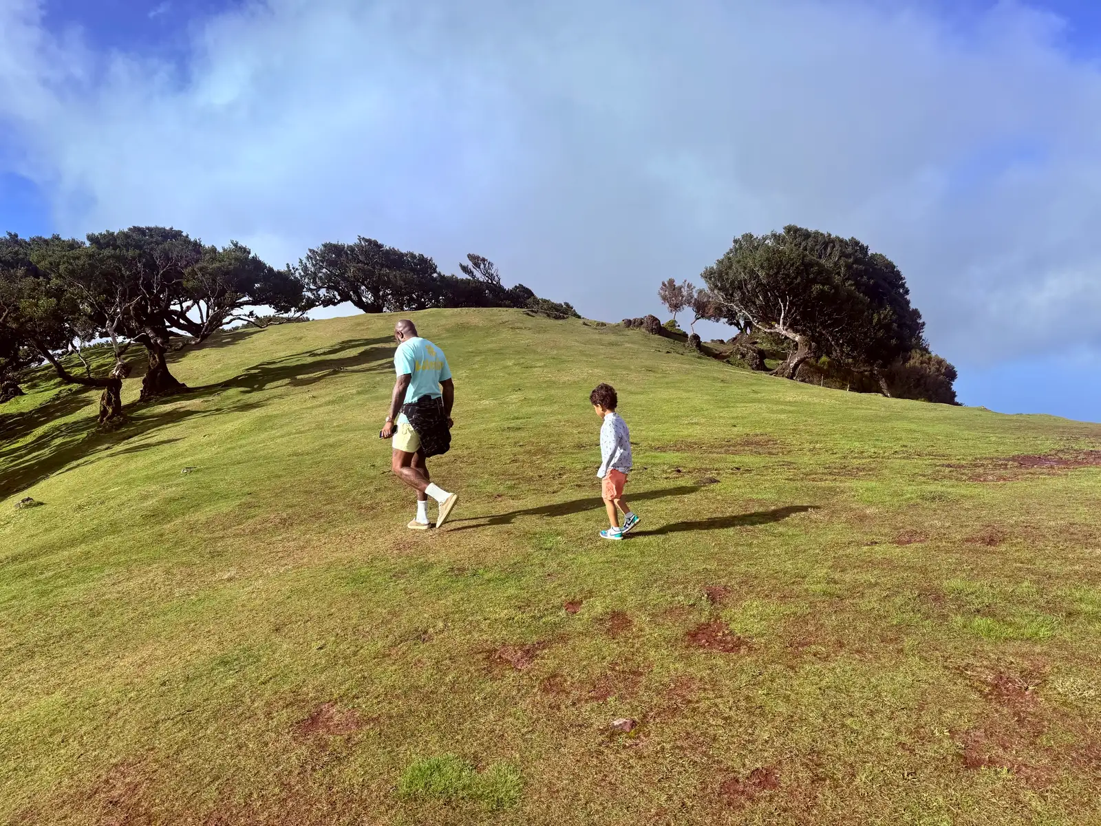 Hugo walking up a grassy ridge with one of his children, wind-bent trees and open sky ahead